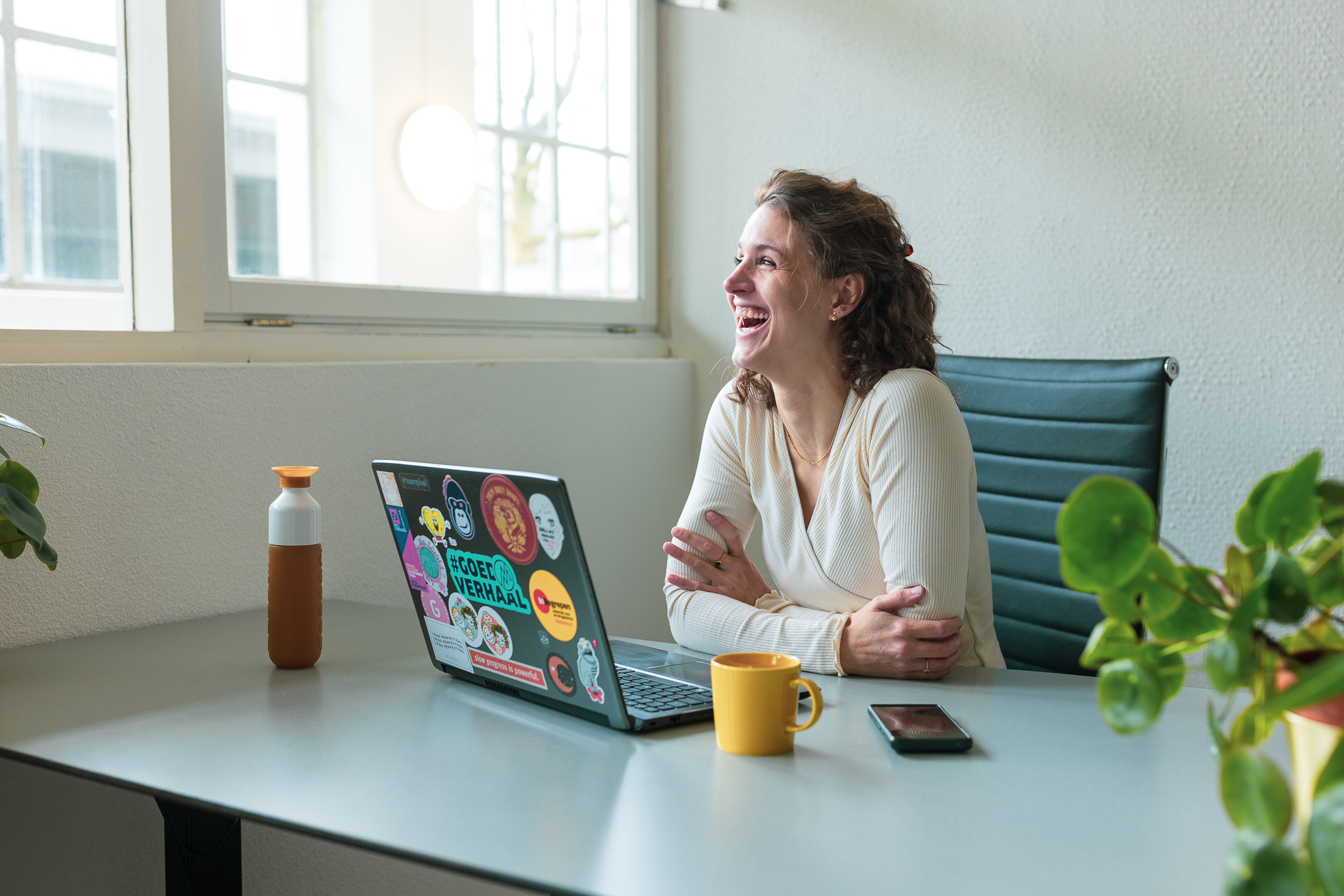 Lisa zit lachend achter haar bureau, op het bureau staat een laptop, flesje water, kop koffie, mobiel en een plantje.